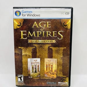 Puede incluir: Caja de CD para PC del juego "Age of Empires Gold Edition". La portada es marrón con letras y detalles dorados. El título del juego se muestra de forma destacada, con imágenes más pequeñas del contenido del juego. La caja incluye el logotipo "Games for Windows".