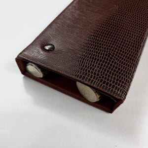 May include: A brown, rectangular case with a textured, reptile-like pattern. The case is closed with a small, dark button. Two silver-colored objects are visible inside the open end of the case.
