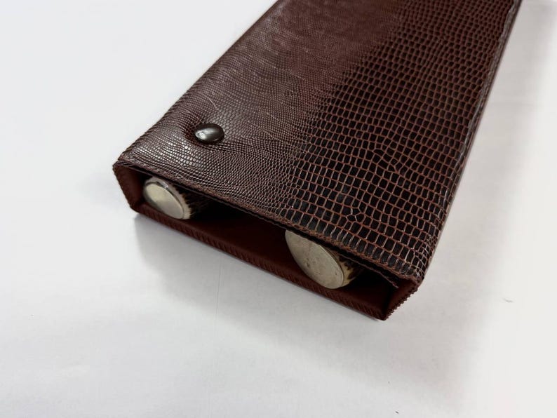 May include: A brown, rectangular case with a textured, reptile-like pattern. The case is closed with a small, dark button. Two silver-colored objects are visible inside the open end of the case.