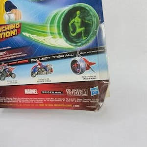 May include: A packaged Spider-Man toy featuring a green figure inside a green and white tube, with a green energy trail. The packaging includes the Marvel and Spider-Man logos, and the text "COLLECT THEM ALL!"