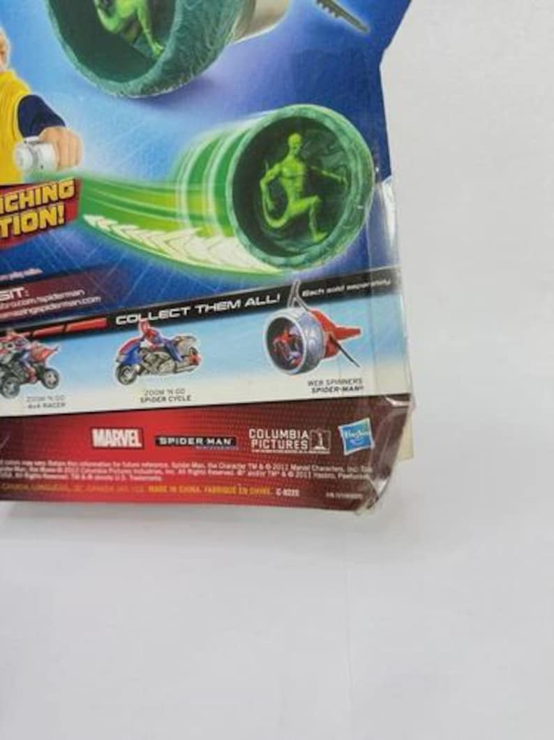 May include: A packaged Spider-Man toy featuring a green figure inside a green and white tube, with a green energy trail. The packaging includes the Marvel and Spider-Man logos, and the text "COLLECT THEM ALL!"