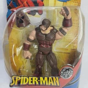 May include: Juggernaut action figure from the Spider-Man series, packaged in a clear plastic blister pack. The figure is mostly brown with red accents and a removable helmet. The packaging features the Spider-Man logo and the words "Super Villain."
