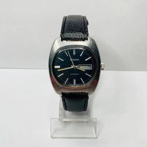 May include: A vintage Bulova automatic wristwatch with a black dial and a black leather band. The watch has a silver-toned case and displays the day and date. The watch is displayed on a clear stand.