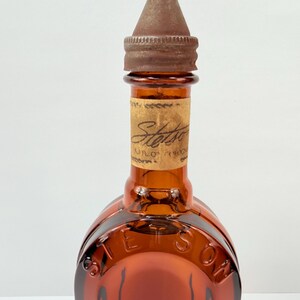 Puede incluir: Botella de vidrio color &aacute;mbar con tapa de metal marr&oacute;n. La botella tiene una etiqueta con la palabra "Stetson" y la misma palabra en relieve en el vidrio. La botella est&aacute; vac&iacute;a.