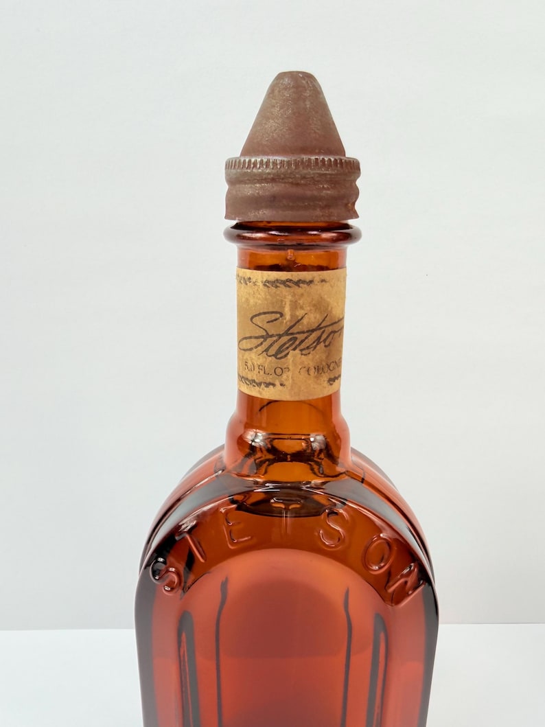Puede incluir: Botella de vidrio color &aacute;mbar con tapa de metal marr&oacute;n. La botella tiene una etiqueta con la palabra "Stetson" y la misma palabra en relieve en el vidrio. La botella est&aacute; vac&iacute;a.