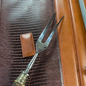 May include: A stainless steel carving fork with a natural antler handle, resting on a brown leather surface. A small, rectangular leather piece and a sharpening steel are also visible. The leather has a reptile-like texture.