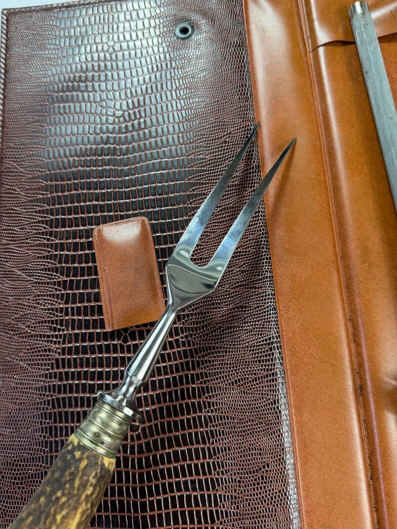 May include: A stainless steel carving fork with a natural antler handle, resting on a brown leather surface. A small, rectangular leather piece and a sharpening steel are also visible. The leather has a reptile-like texture.