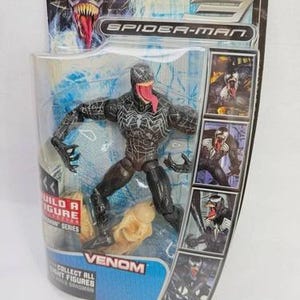 May include: Venom action figure from the Spider-Man series, packaged in a clear plastic blister pack. The figure is black with white spider-web detailing and a long red tongue. The packaging includes the text "VENOM" and "COLLECT ALL RIGHT FIGURES".