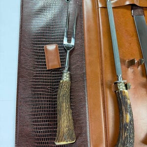 May include: A brown leather case with a reptile skin texture, open to reveal a carving fork, a sharpening steel, and a knife. The handles of the utensils have a rustic, antler-like design.