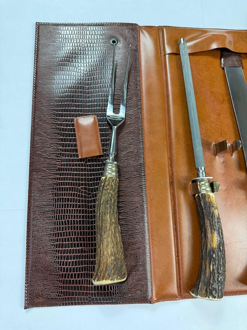 May include: A brown leather case with a reptile skin texture, open to reveal a carving fork, a sharpening steel, and a knife. The handles of the utensils have a rustic, antler-like design.