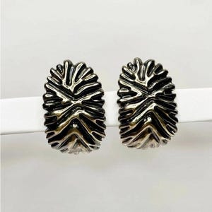 以下が含まれることがあります： シルバーカラーの楕円形スタッドピアス2つ。各イヤリングは、暗いアクセントを持つテクスチャード加工されたリブのデザインが特徴で、印象的なコントラストを生み出しています。イヤリングは白い表面に表示され、金属的な輝きを強調しています。