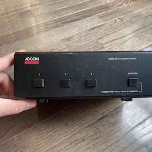 May include: A black ADCOM GFS-3 speaker selector with four square buttons and white text. The text includes "ADCOM", "model GFS-3 speaker selector", "protection", and "engage when two or more are selected". The device is held by a hand.