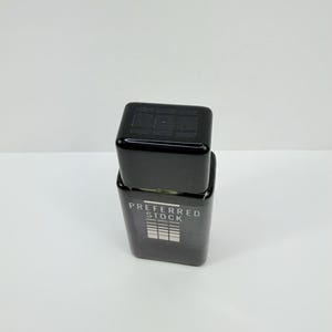 Pu&ograve; includere: Un flacone di profumo rettangolare nero con un tappo abbinato. Il flacone ha le parole "PREFERRED STOCK" stampate sul davanti, insieme a una grafica di linee orizzontali. Il flacone &egrave; appoggiato su una superficie bianca.