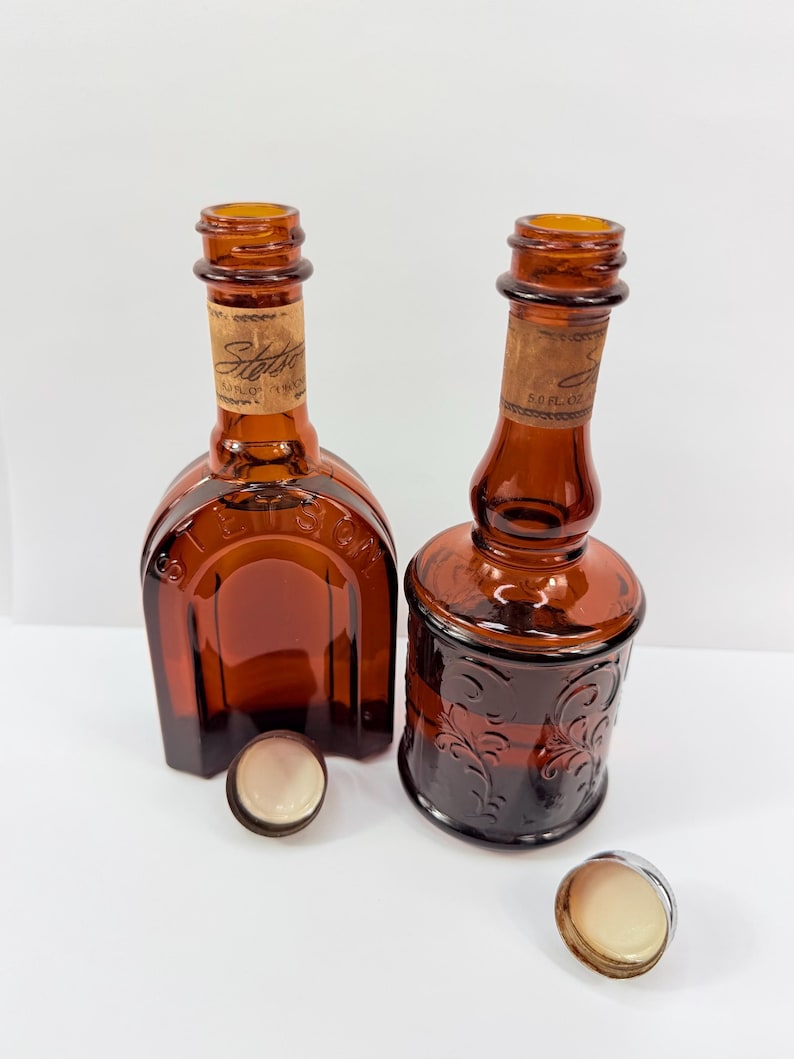 Puede incluir: Dos botellas de vidrio color &aacute;mbar con tapones de corcho y tapas met&aacute;licas. Una botella es cuadrada, la otra redonda con un dise&ntilde;o floral en relieve. Las botellas tienen etiquetas que dicen "Stetson". Las tapas est&aacute;n a un lado.