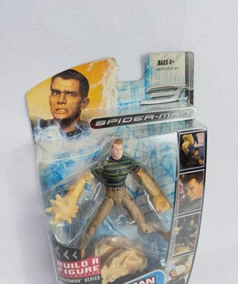 May include: Spider-Man Sandman action figure in original packaging. The figure has a green and black striped shirt, tan trousers, and sand-like arms. The packaging includes the text "Spider-Man" and "Build a Figure".