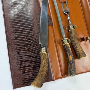 May include: A vintage steak knife set with a brown leather case. The set includes a large knife, a sharpening steel, and a fork. The handles are made of a light brown material with a textured appearance. The case is brown with a reptile-like pattern.