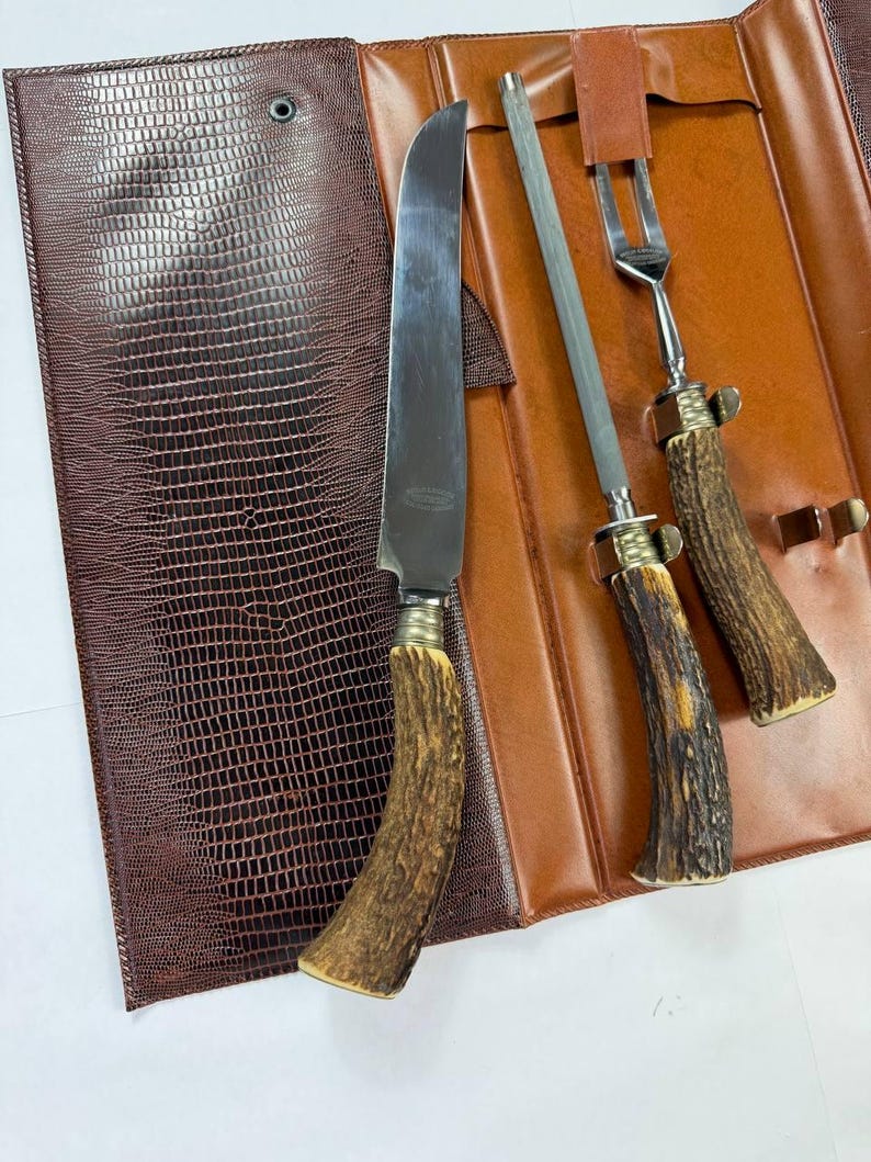 May include: A vintage steak knife set with a brown leather case. The set includes a large knife, a sharpening steel, and a fork. The handles are made of a light brown material with a textured appearance. The case is brown with a reptile-like pattern.