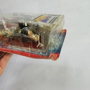 Peut inclure: Une figurine d'action emball&eacute;e, enferm&eacute;e dans du plastique transparent. La figurine est partiellement visible, avec une t&ecirc;te sombre et un corps de couleur beige. L'emballage a une bordure rouge et bleue avec du texte.