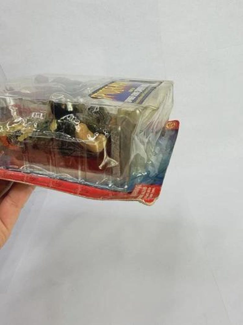 Peut inclure: Une figurine d'action emball&eacute;e, enferm&eacute;e dans du plastique transparent. La figurine est partiellement visible, avec une t&ecirc;te sombre et un corps de couleur beige. L'emballage a une bordure rouge et bleue avec du texte.