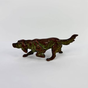 Pode incluir: Estatueta de cão em bronze com pátina verde. O cão está numa pose de corrida, com a cauda levantada e as patas dianteiras estendidas. A estatueta é detalhada e parece ser feita de metal.