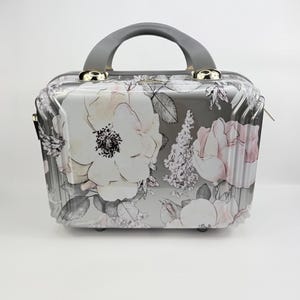 May include: A hard-shell vanity case featuring a gray background with a floral print of large white and pale pink flowers, green leaves, and dark gray foliage. The case has a gray top handle, metallic hardware, and a structured rectangular shape.