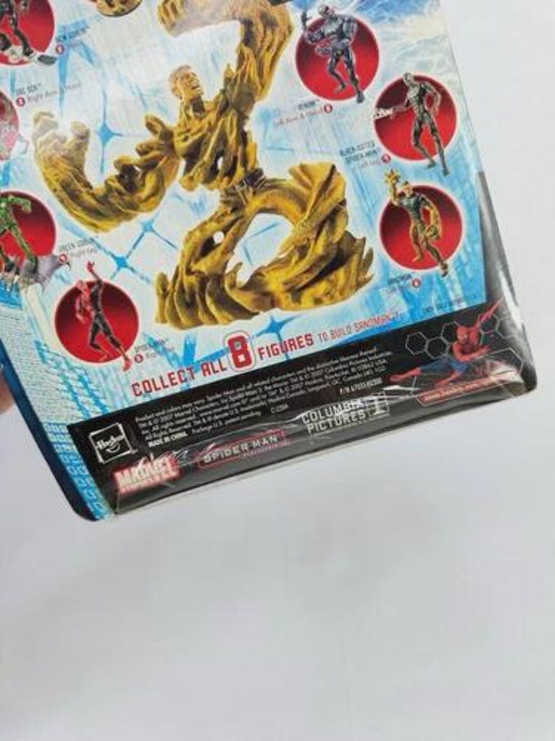 May include: A packaged action figure featuring a large, tan-colored Sandman figure. The packaging includes images of other Spider-Man characters in red circles. Text on the packaging reads "COLLECT ALL 8 FIGURES TO BUILD SANDMAN". The Marvel and Spider-Man logos are visible.