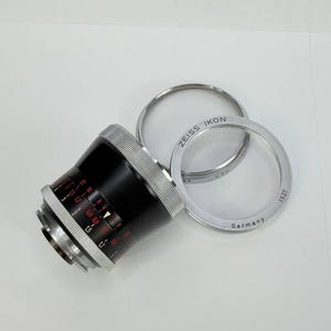 Puede incluir: Un objetivo de c&aacute;mara negro y plateado con marcas rojas y blancas, y dos anillos plateados. Los anillos est&aacute;n marcados "ZEISS IKON" y "Germany". El objetivo tiene una serie de n&uacute;meros y medidas.