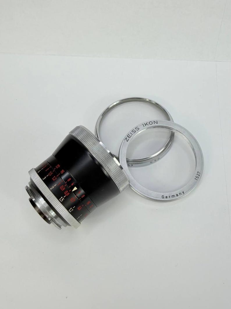 Puede incluir: Un objetivo de c&aacute;mara negro y plateado con marcas rojas y blancas, y dos anillos plateados. Los anillos est&aacute;n marcados "ZEISS IKON" y "Germany". El objetivo tiene una serie de n&uacute;meros y medidas.