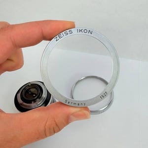 Puede incluir: Un anillo de lente Zeiss Ikon plateado con el texto "ZEISS IKON" y "Germany" grabado. El anillo se sostiene frente a una lente de c&aacute;mara y otro anillo. El n&uacute;mero "1527" tambi&eacute;n est&aacute; grabado en el anillo.