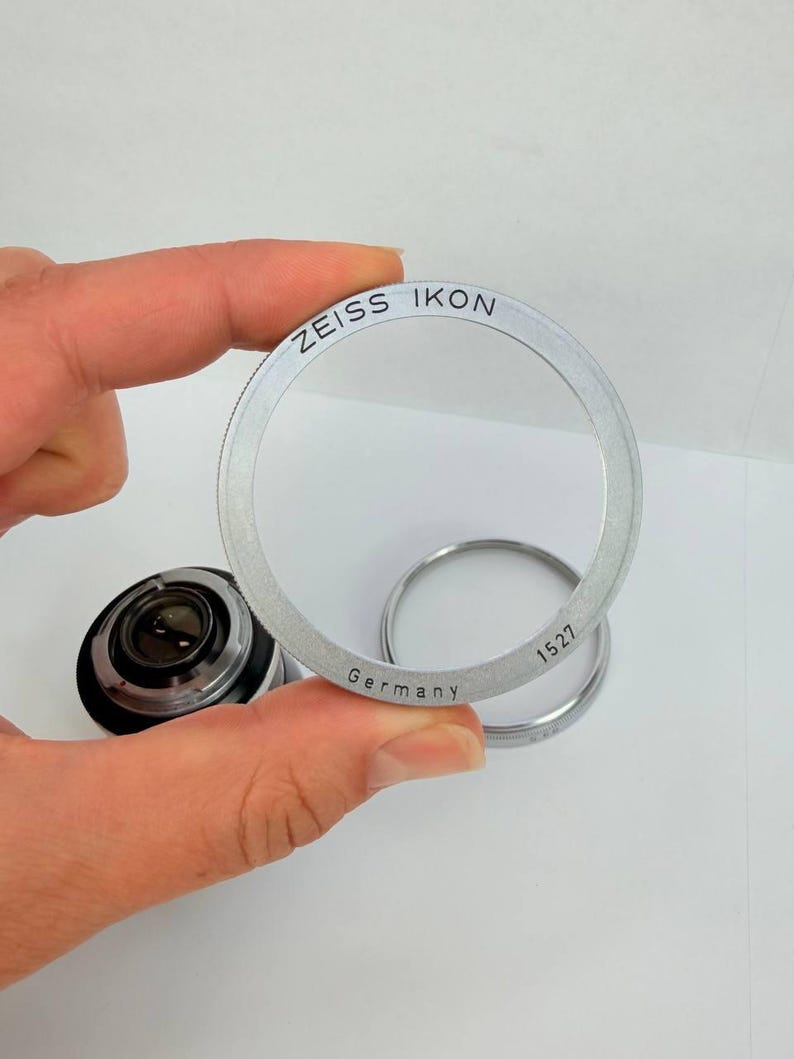 Puede incluir: Un anillo de lente Zeiss Ikon plateado con el texto "ZEISS IKON" y "Germany" grabado. El anillo se sostiene frente a una lente de c&aacute;mara y otro anillo. El n&uacute;mero "1527" tambi&eacute;n est&aacute; grabado en el anillo.
