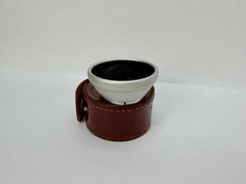Puede incluir: Un parasol plateado con interior negro descansa sobre un estuche de cuero marr&oacute;n. El parasol tiene texto alrededor del borde. El estuche es redondo, con un dise&ntilde;o cosido y un peque&ntilde;o cierre.
