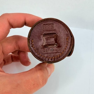 Puede incluir: Objeto circular de cuero marr&oacute;n con las inscripciones "778" y "Germany". El objeto se sostiene en una mano sobre un fondo blanco. El cuero tiene un borde cosido.