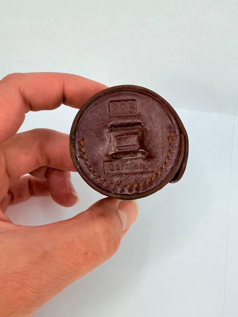Puede incluir: Objeto circular de cuero marr&oacute;n con las inscripciones "778" y "Germany". El objeto se sostiene en una mano sobre un fondo blanco. El cuero tiene un borde cosido.