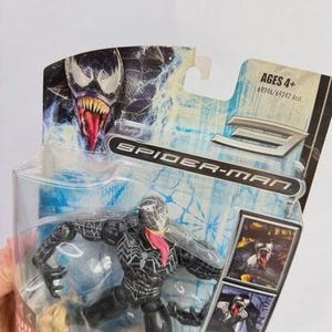 May include: A Venom action figure from the Spider-Man series, packaged in a clear plastic case. The figure is black with white accents and a long pink tongue. The packaging features the Spider-Man logo and the text "AGES 4+".