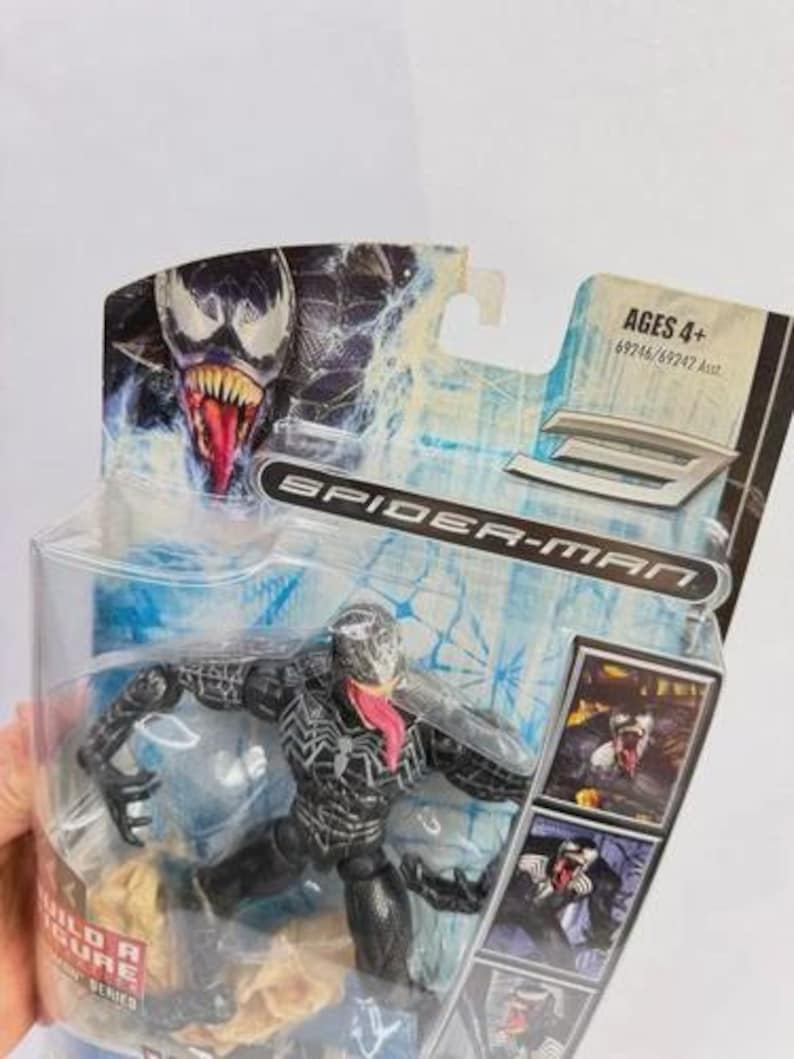 May include: A Venom action figure from the Spider-Man series, packaged in a clear plastic case. The figure is black with white accents and a long pink tongue. The packaging features the Spider-Man logo and the text "AGES 4+".