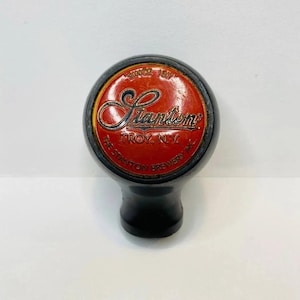 Puede incluir: Un tirador de grifo negro y rojo de Stanton Brewery. La parte superior es redonda con el nombre "Stanton" en letras doradas sobre un fondo rojo. También se ve el texto "TROY, N.Y." y "THE STANTON BREWERY INC.".