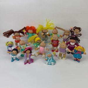 Może przedstawiać: Kolekcja piętnastu małych plastikowych lalek Cabbage Patch Kids z włosami z włóczki, z których każda ma na sobie inny kolorowy strój. Figurki przyjmują różne pozy, w tym stojące i siedzące, a niektóre trzymają małe akcesoria.