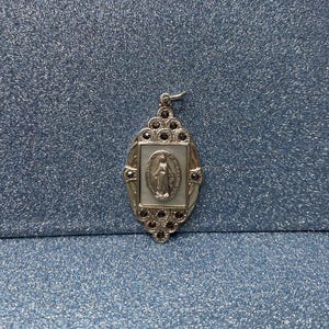May include: Silver-toned pendant with an oval frame, featuring a central image of a religious figure. The pendant is adorned with small, dark-colored embellishments around the frame and a bail at the top for hanging. The background is a textured, blue surface.