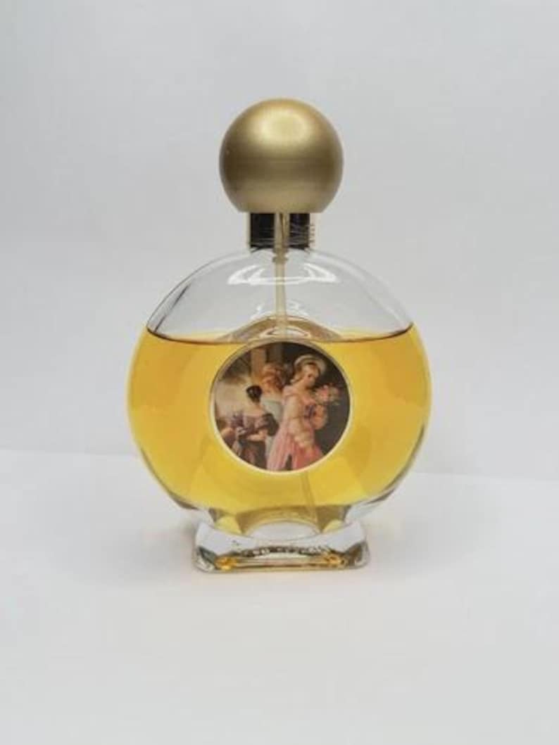 May include: A clear, round perfume bottle with a gold-colored spherical cap. The bottle contains a yellow liquid and features a circular image of figures in a classical painting. The base of the bottle is clear and rectangular.
