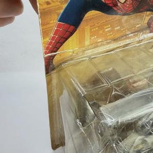 May include: Spider-Man action figure in original packaging. The figure is in a crouching pose, with a red and blue costume. The packaging is yellowed and shows signs of wear. The figure is in a clear plastic case.
