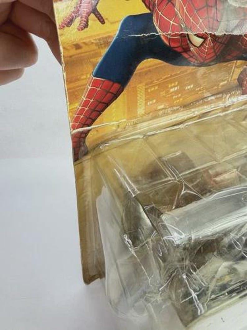 May include: Spider-Man action figure in original packaging. The figure is in a crouching pose, with a red and blue costume. The packaging is yellowed and shows signs of wear. The figure is in a clear plastic case.
