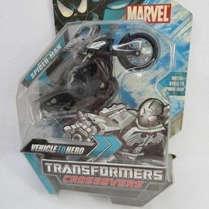 Puede incluir: Un juguete de moto de Spider-Man negro en un paquete de pl&aacute;stico transparente. El paquete presenta el logotipo de Marvel y el texto "Transformers Crossovers". El juguete est&aacute; etiquetado como "Vehicle to Hero".