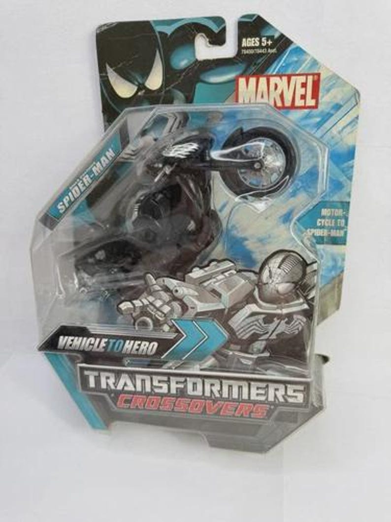 Puede incluir: Un juguete de moto de Spider-Man negro en un paquete de pl&aacute;stico transparente. El paquete presenta el logotipo de Marvel y el texto "Transformers Crossovers". El juguete est&aacute; etiquetado como "Vehicle to Hero".