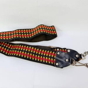 May include: A black camera strap with a repeating pattern of red and yellow rectangles. The strap has black leather ends with metal rivets and silver-colored metal clips for attachment.