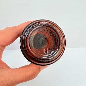 Puede incluir: Un tarro de vidrio &aacute;mbar oscuro, visto desde abajo, con forma circular y superficie texturizada. El tarro tiene una base ligeramente redondeada y un acabado oscuro y brillante. El n&uacute;mero "13" est&aacute; grabado en la parte inferior.