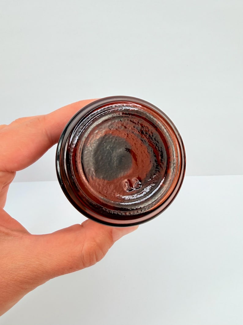 Puede incluir: Un tarro de vidrio &aacute;mbar oscuro, visto desde abajo, con forma circular y superficie texturizada. El tarro tiene una base ligeramente redondeada y un acabado oscuro y brillante. El n&uacute;mero "13" est&aacute; grabado en la parte inferior.