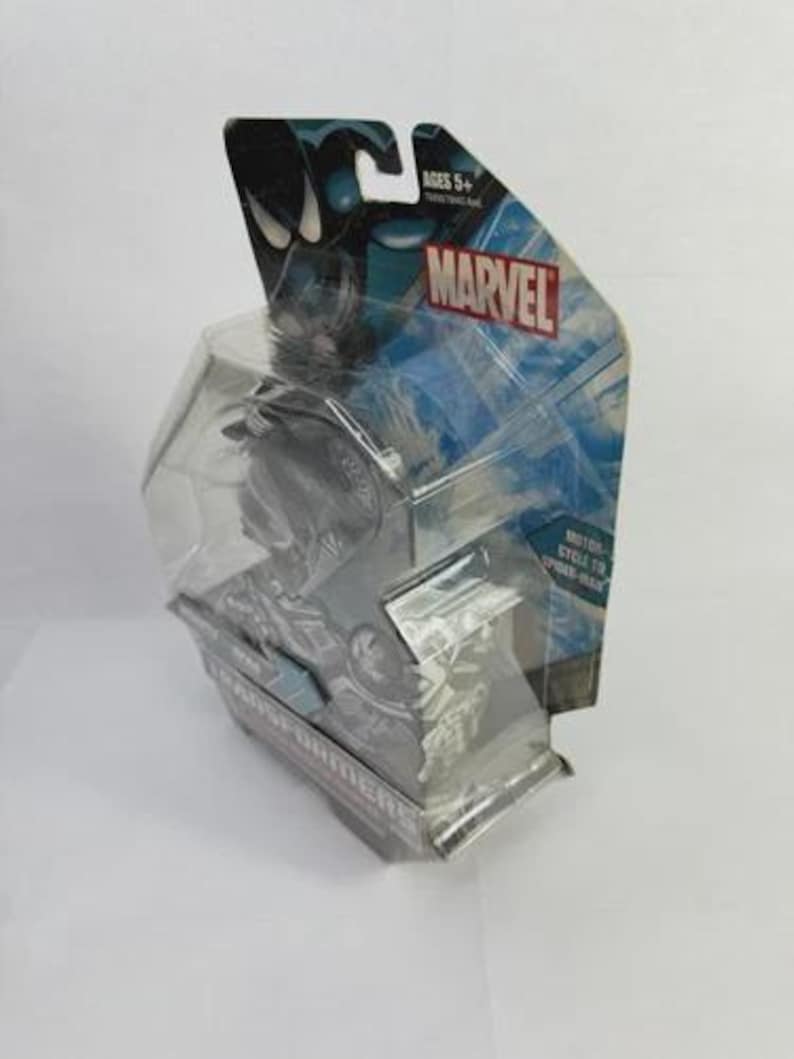 Puede incluir: Una figura de acci&oacute;n Marvel empaquetada, con un personaje negro y gris. El embalaje incluye el logotipo de Marvel, el texto "Ages 5+" y las palabras "Motor Cycle to Spider-Man". La figura est&aacute; encerrada en una carcasa de pl&aacute;stico transparente.