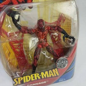 May include: A red and black Carnage action figure from the Spider-Man series, in a clear plastic blister pack. The figure is posed with outstretched arms and has detailed features. The packaging features the Spider-Man logo and the words "Super Villain."