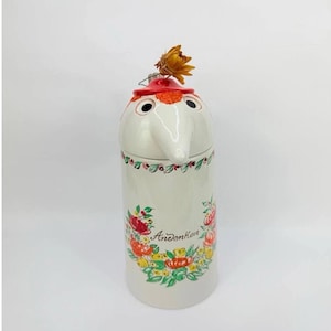 May include: A cylindrical ceramic lidded jar with a cream-colored glaze, featuring a hand-painted floral wreath in red, orange, yellow, and green. The word "Andenken" is written in black script. A small dried orange flower sits on the red lid.