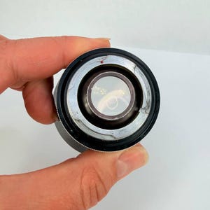 Puede incluir: Primer plano de una lente de c&aacute;mara, con un anillo de metal plateado y un agarre de goma negro. La lente tiene un elemento de vidrio transparente en el centro. Un peque&ntilde;o punto rojo es visible en el anillo de metal.